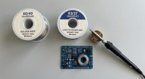 A clean, top-down shot of two solder spools side by side — one labelled "60/40 (Sn60Pb40)" and the other "63/37 (Sn63Pb37)" — with a soldering iron and a small PCB in the frame. Keep the background plain so the labels read clearly.